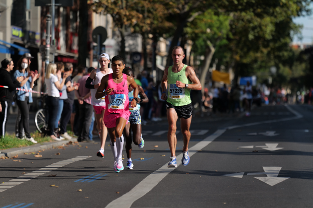 Gruppe von Läufern bei einem Stadtmarathon mit Zuschauern auf der linken Seite, unscharfer städtischer Hintergrund mit Bäumen, Gebäuden und einem Fahrrad.