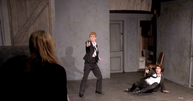 Zwei Frauen in schwarzen Anzügen, eine sitzend auf dem Boden und die andere stehend, beide mit Waffen, mit einer dritten Frau in der Nähe, in einem Raum mit einem Stuhl, einer Toilette und einer hölzernen Tür.