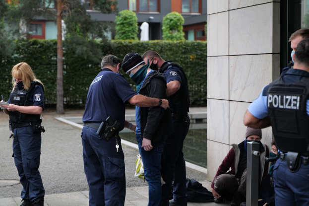 Eine Gruppe von Polizisten steht neben einem Gebäude, während eine Frau ein Buch und einen Stift hält, mit Menschen in der Nähe und Grün im Hintergrund.