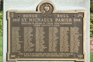 Eine Gedenktafel an einer Steinwand in der St. Michael's Parish, mit eingravierter Inschrift, umgeben von grünem Gras und Bäumen.