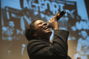 Eine Frau in einem schwarzen Kleid mit einem Dutt singt leidenschaftlich in ein Mikrofon auf der Bühne, ihr Gesicht von hellen Scheinwerfern beleuchtet.