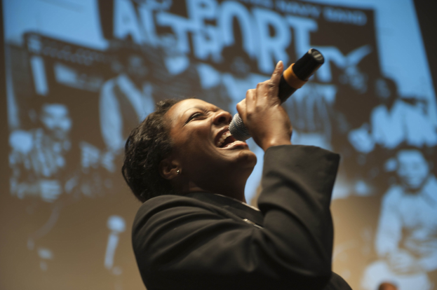 Eine Frau in einem schwarzen Kleid mit einem Dutt singt leidenschaftlich in ein Mikrofon auf der Bühne, ihr Gesicht von hellen Scheinwerfern beleuchtet.