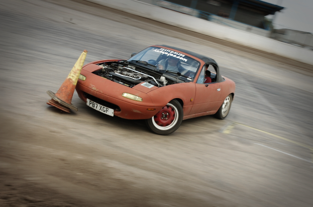 Eine Person fährt ein rotes Auto mit hoher Geschwindigkeit auf einer Rennstrecke, mit einem Verkehrskegel vor sich.