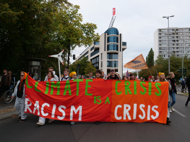 Eine Gruppe von Menschen marschiert eine von Bäumen gesäumte Straße entlang, mit einer Tafel, auf der 'Climate Crisis is a Crisis' steht, im Hintergrund Gebäude und einen klaren Himmel.