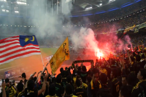 Eine Menschenmenge in einem Stadion mit Bengalos, Fahnen und einer Kamera, mit Rauch und Schildern mit der Aufschrift "Malaysia vs Malaysia Live Streaming" im Hintergrund.