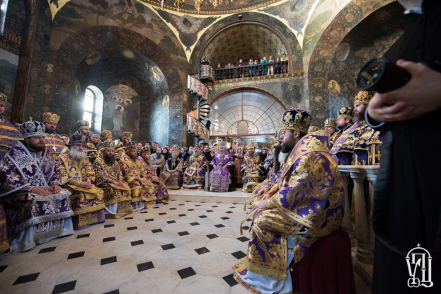 Eine Gruppe von Menschen in einer Kirche, einige sitzen und andere stehen auf einer Treppe, mit einer Person, die rechts eine Kamera hölt; eine Kristallleuchter hängt von der Decke, und Gemälde schmücken die Wände.