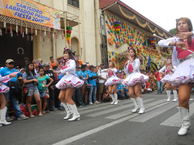 Eine Gruppe von Frauen in rosänen und weißen Outfits tanzt auf der Straße, umgeben von einer Menge von Menschen, die stehen und zuschauen, mit Gebäuden, Bäumen und einem klaren blauen Himmel im Hintergrund.