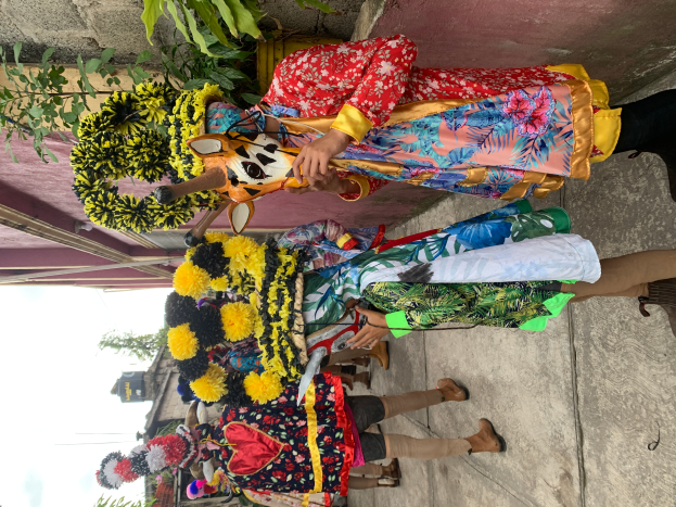 Eine Gruppe von Menschen in farbenfrohen Kostümen steht vor einem Gebäude und hält verschiedene Gegenstände, mit Bäumen, Gebäuden und einem klaren blauen Himmel im Hintergrund während eines Karnevalsumzugs.