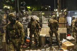 Eine Gruppe von Polizisten in Uniform und Helmen steht in einer Reihe vor einem Zaun bei Nacht, hält Gewehre und hat Müll und Trümmer auf dem Boden verstreut, sowie Gebäude, Bäume und ein Schild im Hintergrund.